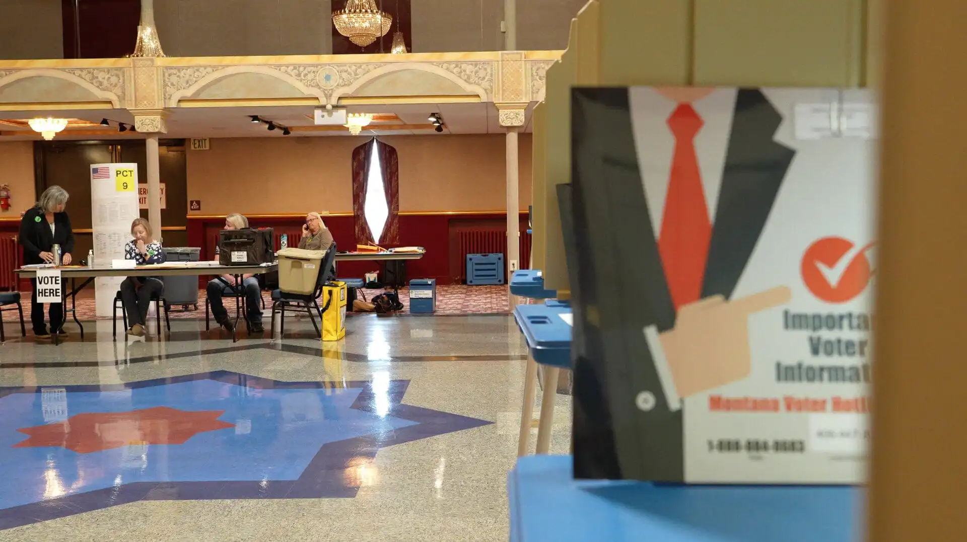 A voting center with poll workers seated at a table in the background and a sign in the foreground displaying voter information with a checkmark and a suit illustration, captured during Montana Election season. Blue voting booths line the right side of the image.